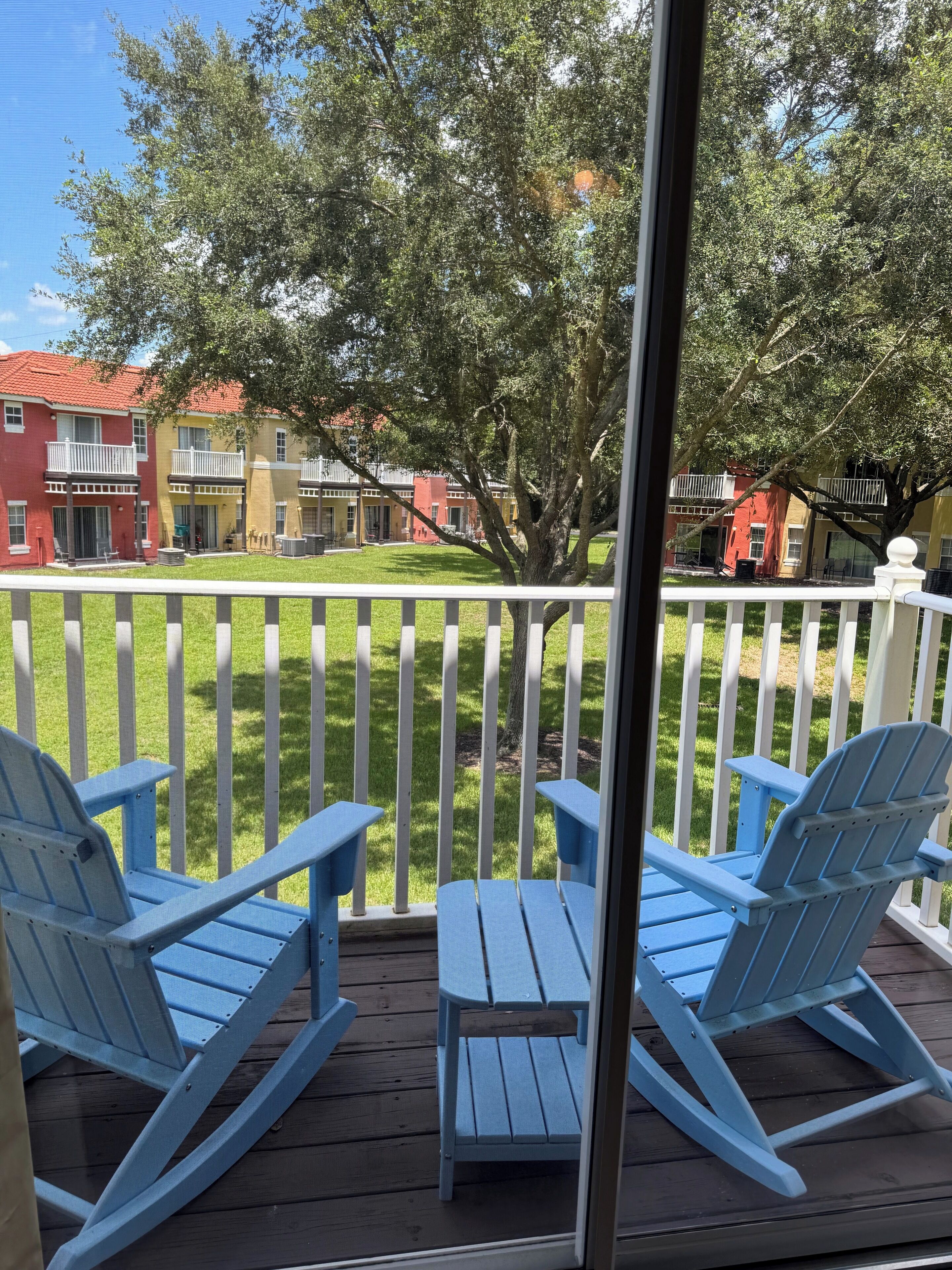Eine Terrasse mit zwei blauen Adirondack-Stühlen und einem kleinen Tisch, mit Blick auf eine Rasenfläche mit weißem Zaun und Gebäuden im Hintergrund.