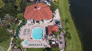 An aerial view of a resort with a large swimming pool, multiple lounge areas, and a red-roofed building.