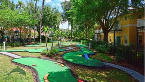 A miniature golf course with green and red obstacles, surrounded by trees and a yellow building.