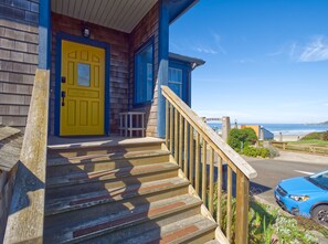 A wooden house with a yellow door and blue trim, a wooden staircase, and a view of the ocean.