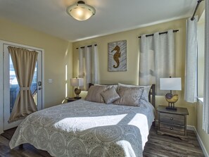A bedroom with a large bed, two bedside tables with lamps, a door with curtains, and a seahorse artwork on the wall.