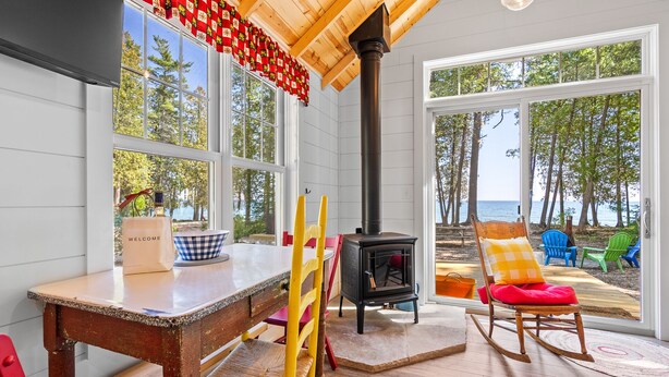 Ein gemütliches Zimmer mit Holztisch, Stühlen, einem schwarzen Holzofen und großen Fenstern mit Blick auf Strand und Bäume.