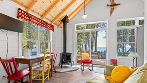 Ein gemütliches Blockhaus-Interieur mit Holzbalkendecke, Kamin, Essbereich mit unterschiedlichen Stühlen und Blick ins Freie.