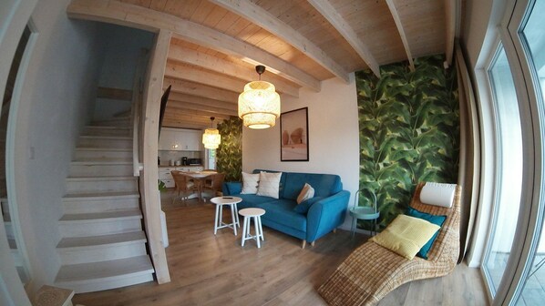 A spacious living area with a blue sofa, a wicker chair, and a dining table. A staircase leads to an upper level.