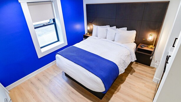 Een moderne slaapkamer met een groot bed, een raam met zonwering, een nachtkastje met lamp en een donkere hoofdbord.