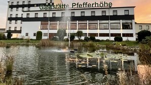 En modern byggnad med en skylt som visar texten ”Gasthof Pfefferhöhe”, placerad framför en damm med näckrosor och en fontän.
