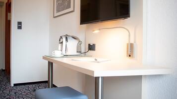A hotel room with a white desk, a flat-screen TV mounted above it, a coffee maker, a cup, and a small stool.