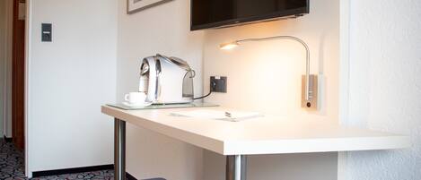 A hotel room with a white desk, a flat-screen TV mounted above it, a coffee maker, a cup, and a small stool.