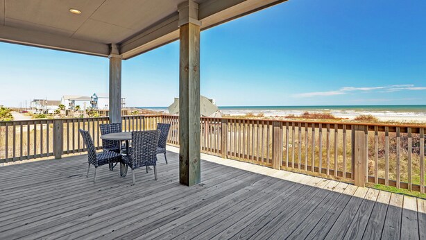 Eine Holzterrasse mit Tisch und Stühlen, mit Blick auf einen Strand und einer Holzbrüstung.