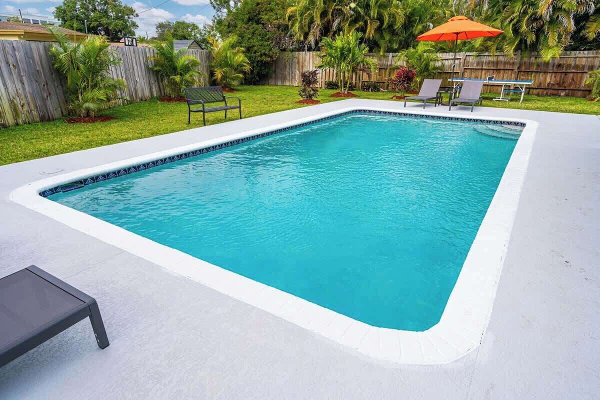Une piscine rectangulaire bordée de blanc, entourée d’une terrasse en béton. On aperçoit des chaises longues et un parasol à l’arrière-plan.