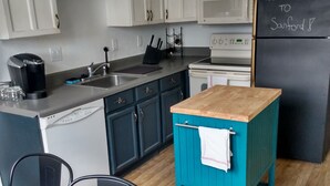 Cozinha moderna com armários brancos, uma geladeira preta com lousa, uma cafeteira e um carrinho verde-água.