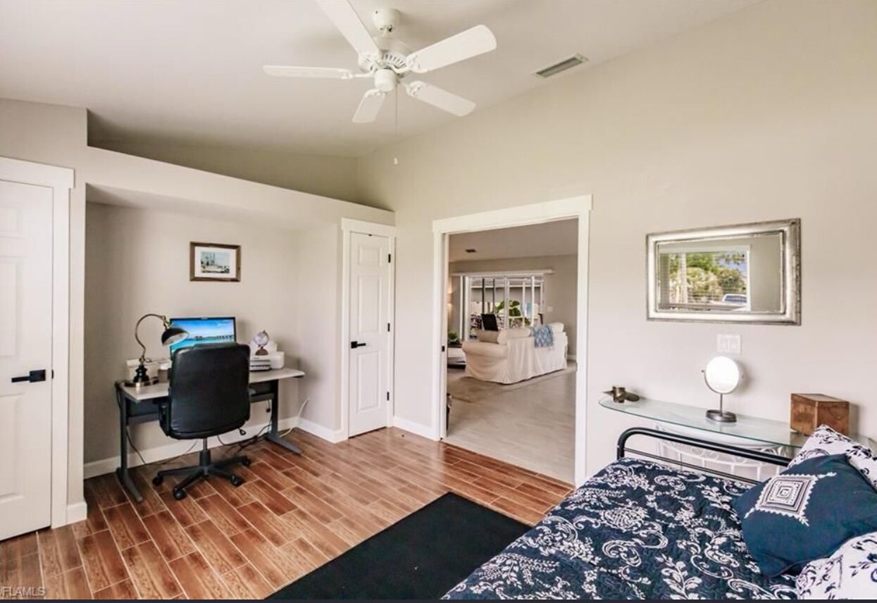 A bedroom with a bed, a desk, a chair, a ceiling fan, and a door leading to another room.