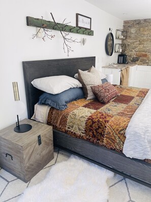 Un dormitorio ordenado con cama, mesita de noche, estante en la pared y pared de piedra.