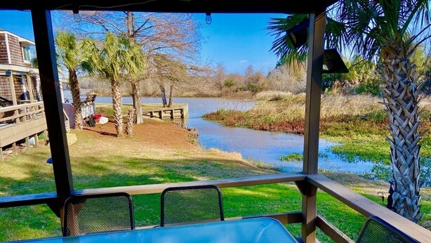 Un espacio al aire libre con techo, con mesa y sillas, que da vista a un cuerpo de agua y una zona verde con árboles.