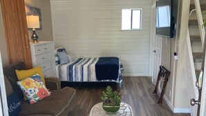 Una sala de estar acogedora con un sofá, una mesita de centro con una planta, una cama con ropa de cama a rayas y un televisor montado en la pared.