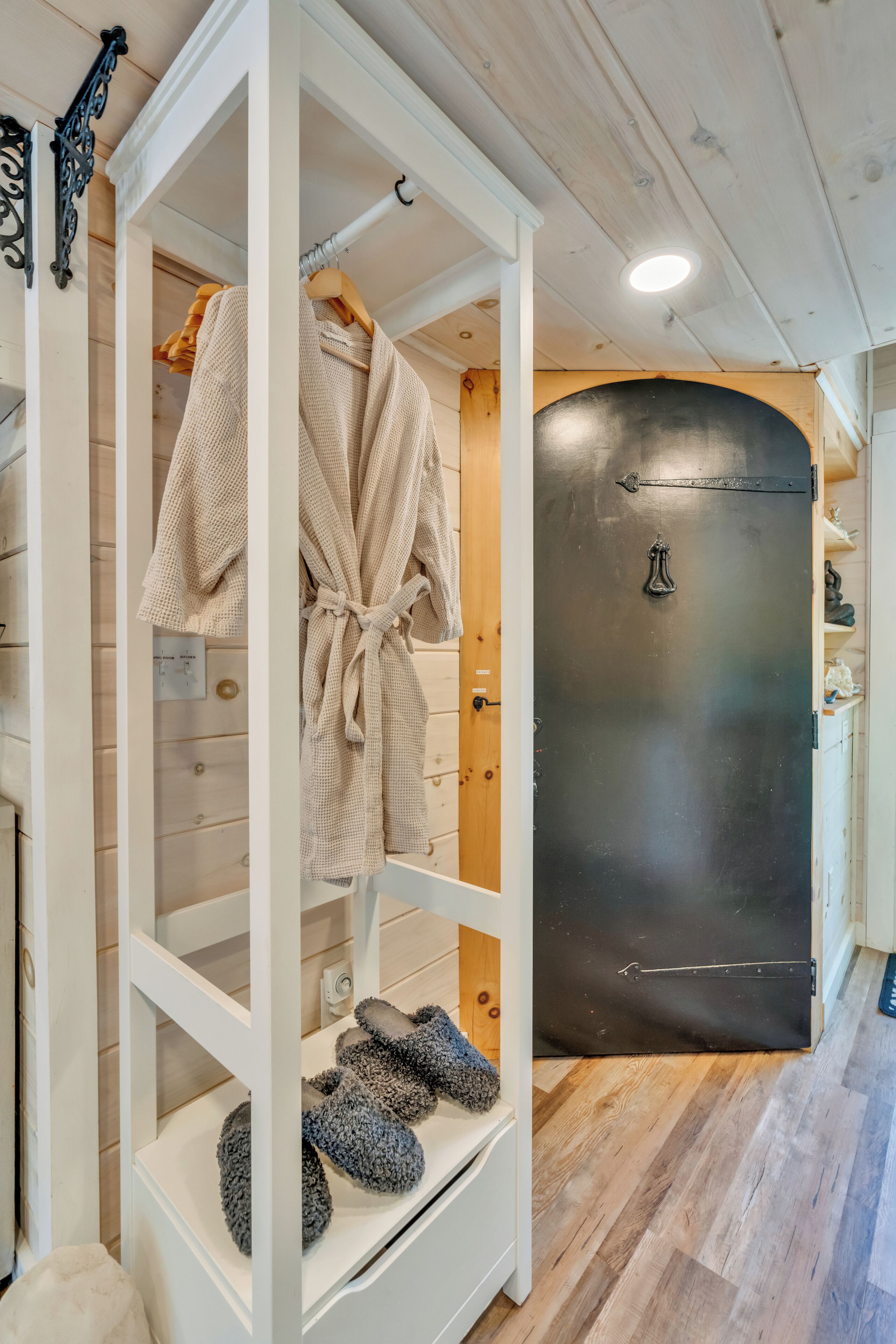 A white wardrobe with a hanging robe and slippers, a black door with a handle, and a wooden floor.