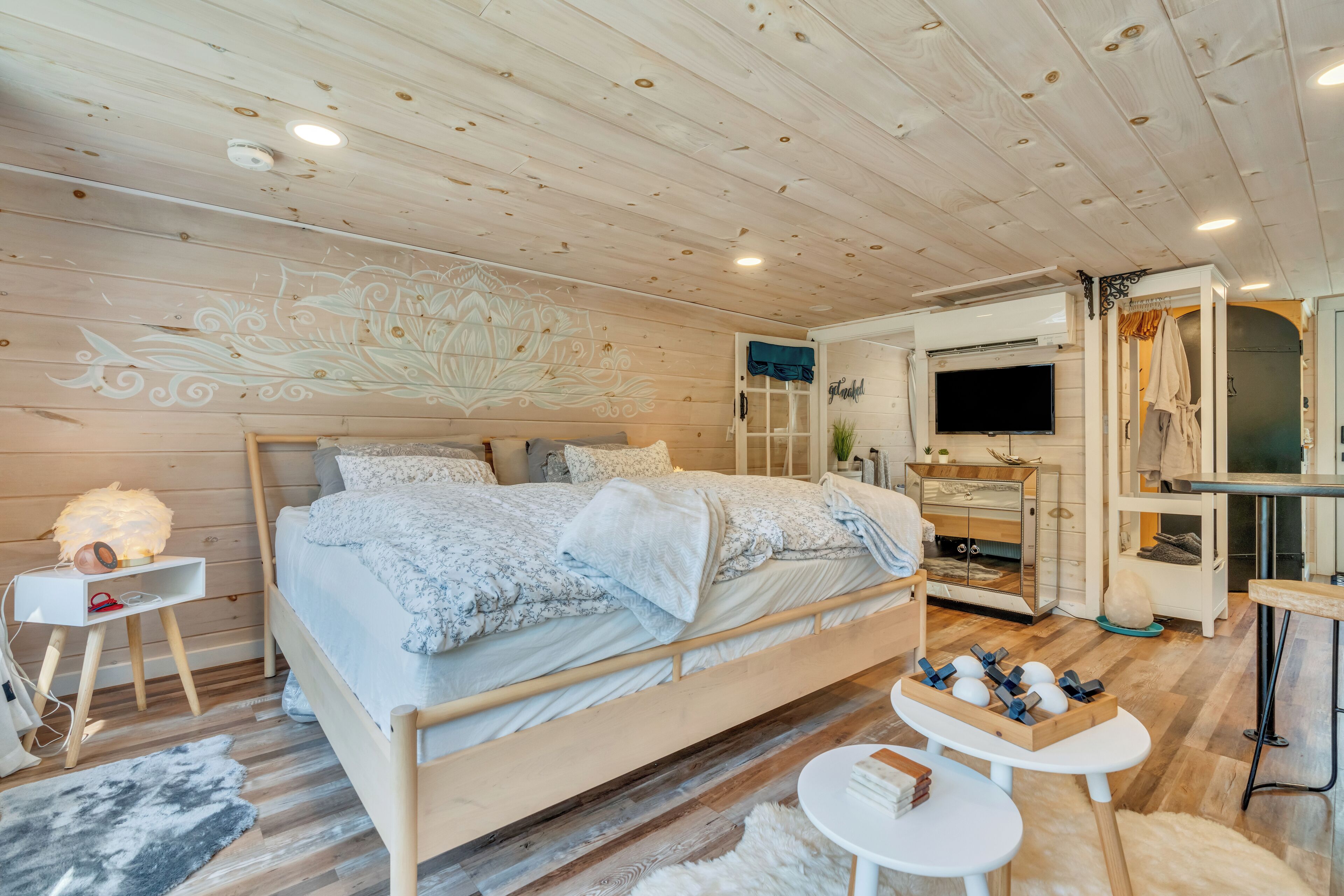 A bedroom with a wooden ceiling, a bed with a floral bedspread, a TV, a vanity with a mirror, and a small table with a basket.