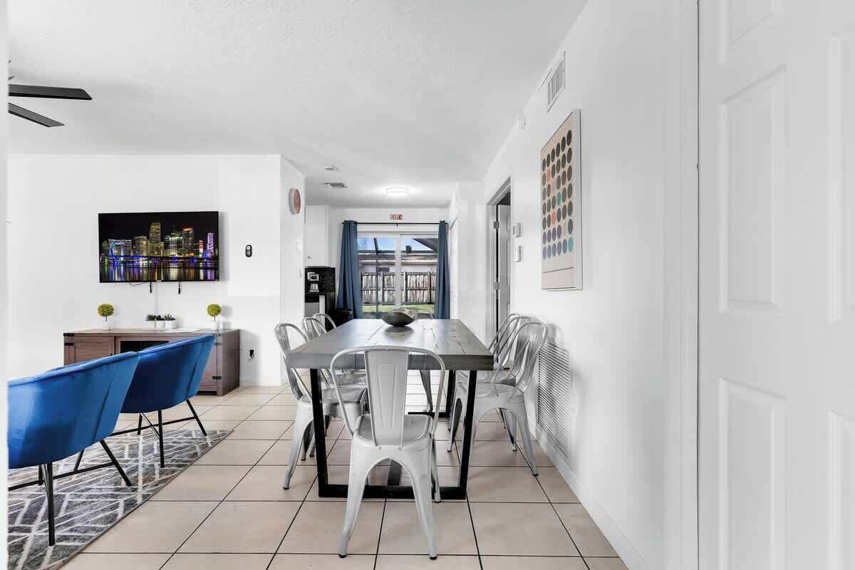 Un espace de salle à manger moderne avec une longue table, des chaises bleues et blanches, et un tableau représentant un paysage urbain accroché au mur.