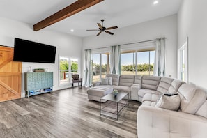 A spacious living room with a sectional sofa, a flat-screen TV, wooden beams, and large windows. - Farm Stay w/ Pool, Animals & Privacy Near Town – Pet Friendly (Millbrook)