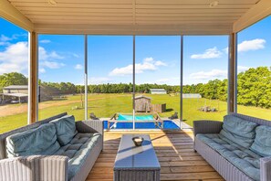 Eine überdachte Terrasse mit Korbmöbeln, ein Pool und Blick auf eine Wiese mit Bäumen.