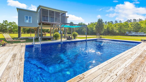 Une piscine avec une terrasse en bois, une maison dotée d’un patio couvert et une cour gazonnée.