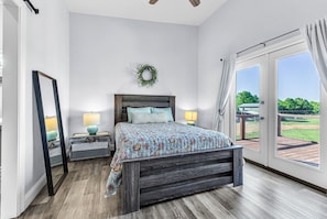 A bedroom with a bed, a nightstand, a mirror, a wall-mounted wreath, and a view of a deck through a sliding glass door. - Farm Stay w/ Pool, Animals & Privacy Near Town – Pet Friendly (Millbrook)