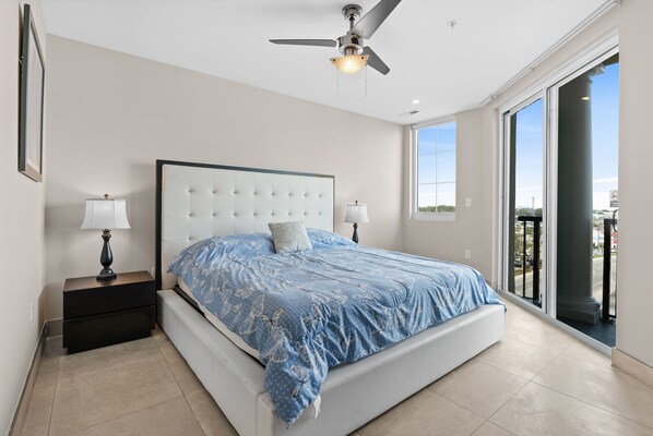 A bedroom with a large bed, two bedside tables with lamps, a ceiling fan, and a view through a large window. - Ocean Pearl: 7 BR Oceanfront Condo (Myrtle Beach)