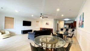 A spacious living room with a glass-top dining table, a sofa, a TV mounted on the wall, and a ceiling fan. - Great 7 BD walk to the beach + pool (North Myrtle Beach)