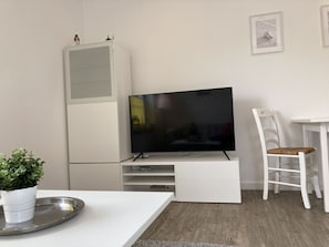 En moderne stue med et fladskærms-tv på et hvidt stativ, et hvidt spisebord med en stol, og en lille potteplante på en bakke.