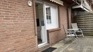 Een bakstenen gebouw met een witte deur, een klein terrasje met een stoel, en een metalen rooster.