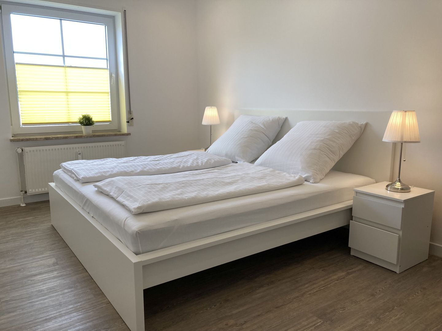 Una cama doble con ropa de cama blanca, dos lámparas de noche, una mesita de noche pequeña y una ventana con persianas.