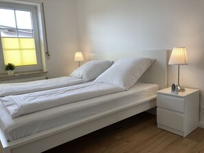 En dobbeltseng med hvide sengetæger, et natbord med lampe, og en lille potteplante på vindueskarmen.