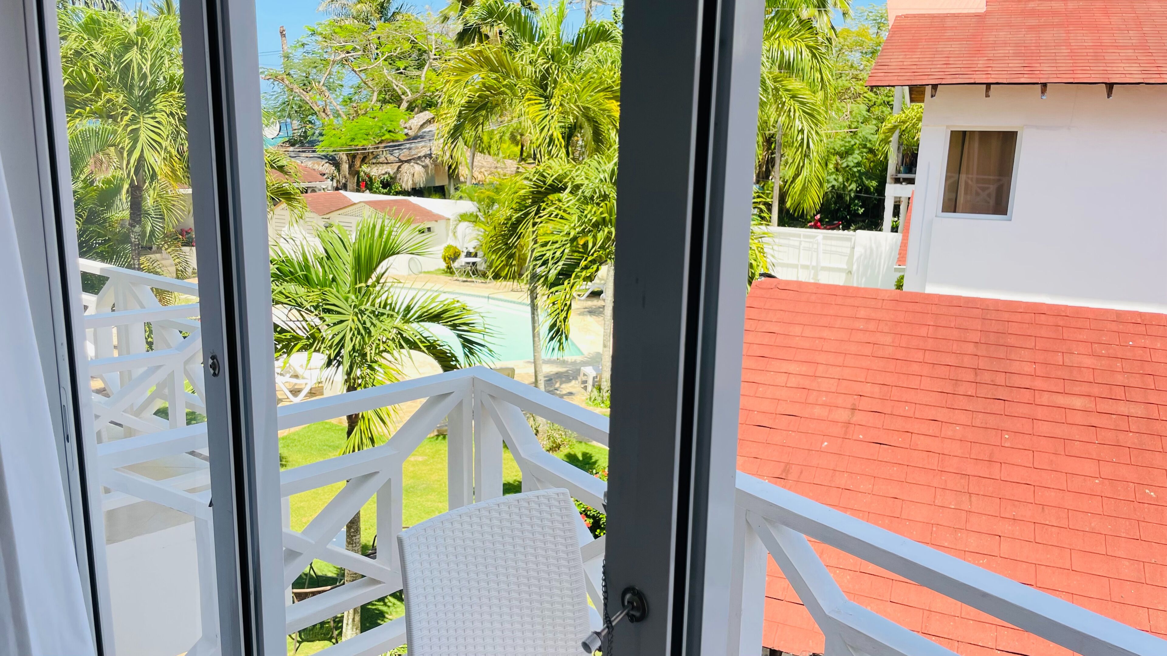Un balcon avec une balustrade blanche, une chaise blanche et une vue sur une piscine, des palmiers et un bâtiment avec un toit rouge.