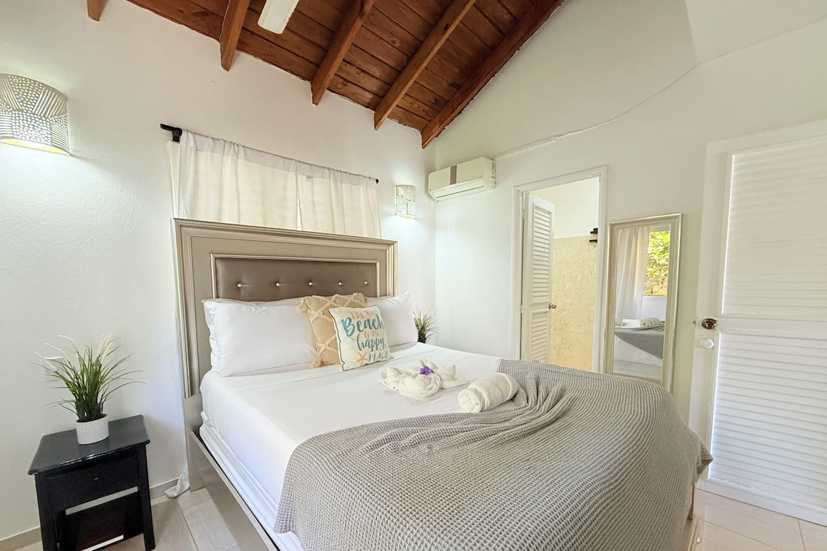 A bedroom with a bed, bedside tables, a nightstand, a window with curtains, an air conditioning unit, and a door.