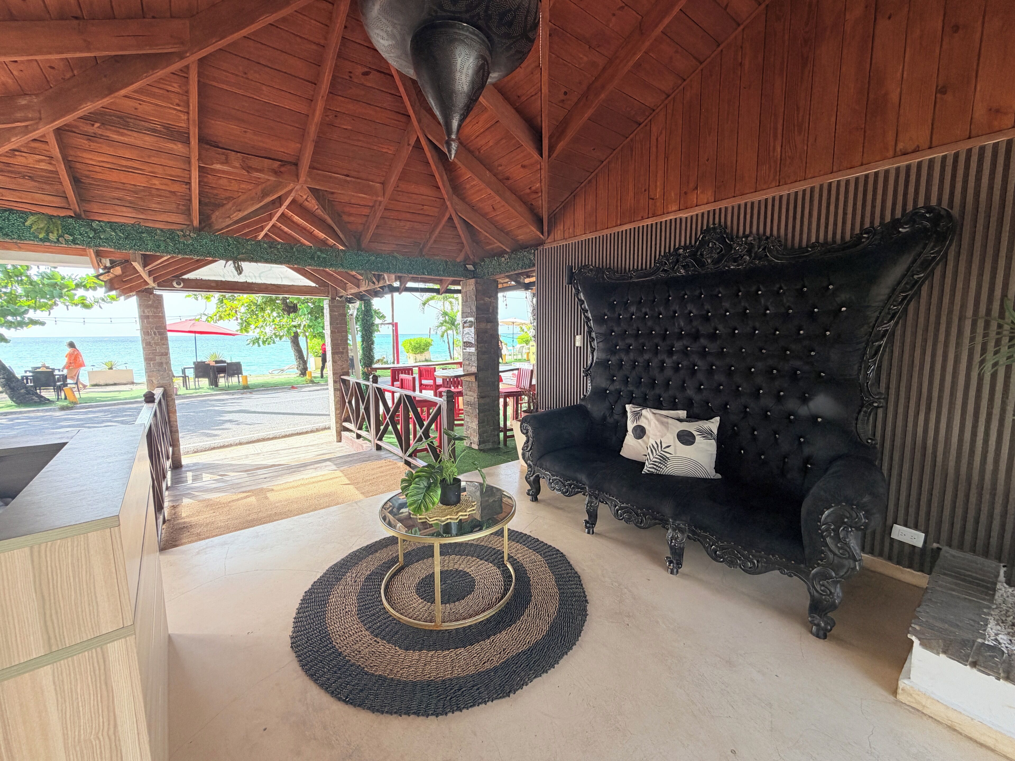 A covered outdoor seating area with a black upholstered sofa, a glass table with a woven rug, and a view of the sea.