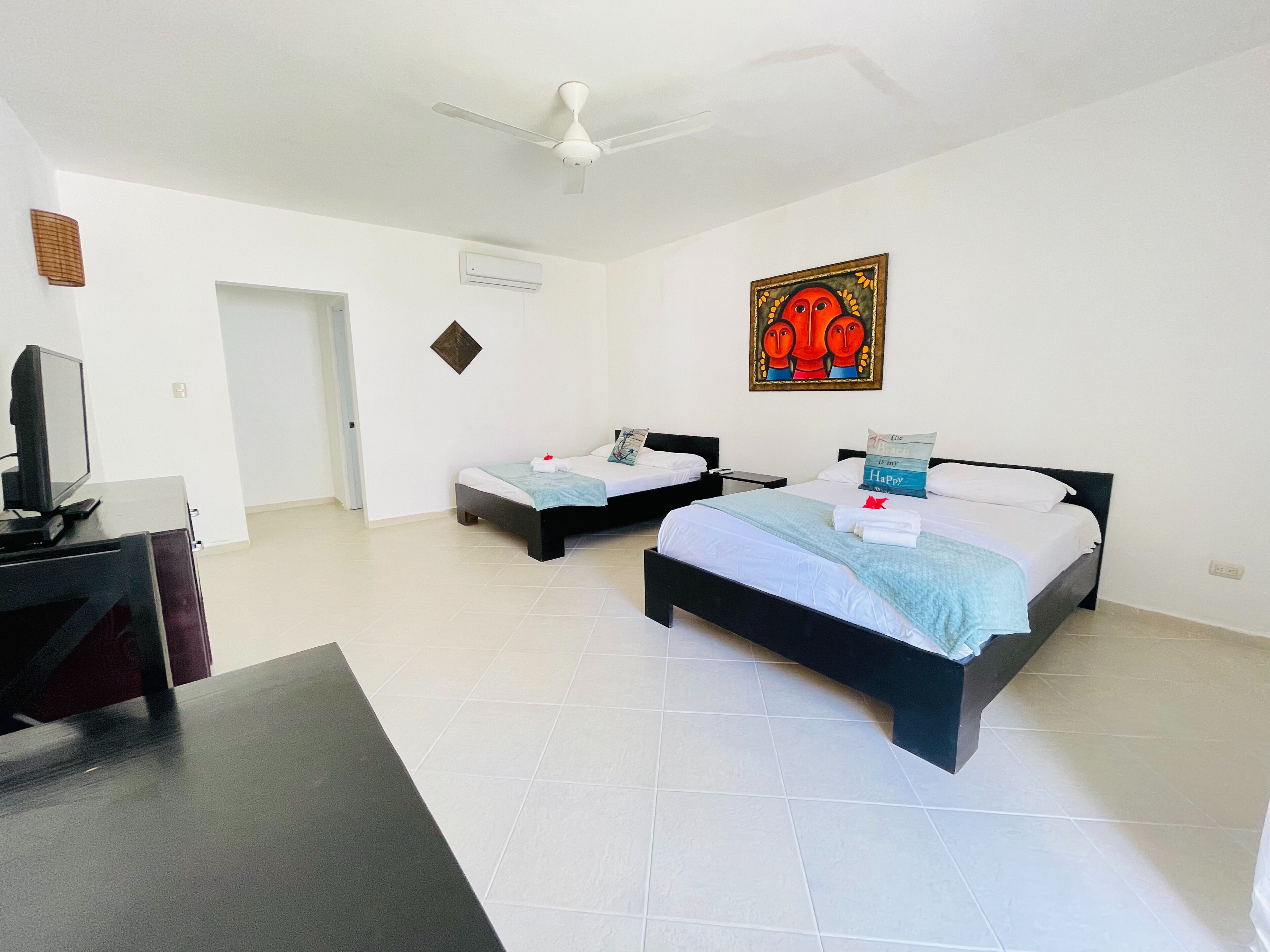 A hotel room with two beds, a desk with a TV, a ceiling fan, and a painting on the wall.