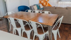 Uma área de refeições com mesa de madeira e cadeiras brancas, um sofá cinza e uma escada ao fundo.