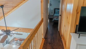Um corredor de madeira estreito com um ventilador de teto, uma escada e uma cama ao fundo.