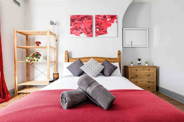 Un lit bien fait, recouvert d’une couverture rouge, une table de chevet en bois avec un miroir, et une étagère en bois ornée de plantes.
