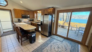 Cocina con armarios de madera, un refrigerador de acero inoxidable, estufa y microondas. Hay un ventanal con vista a una terraza y un cuerpo de agua.