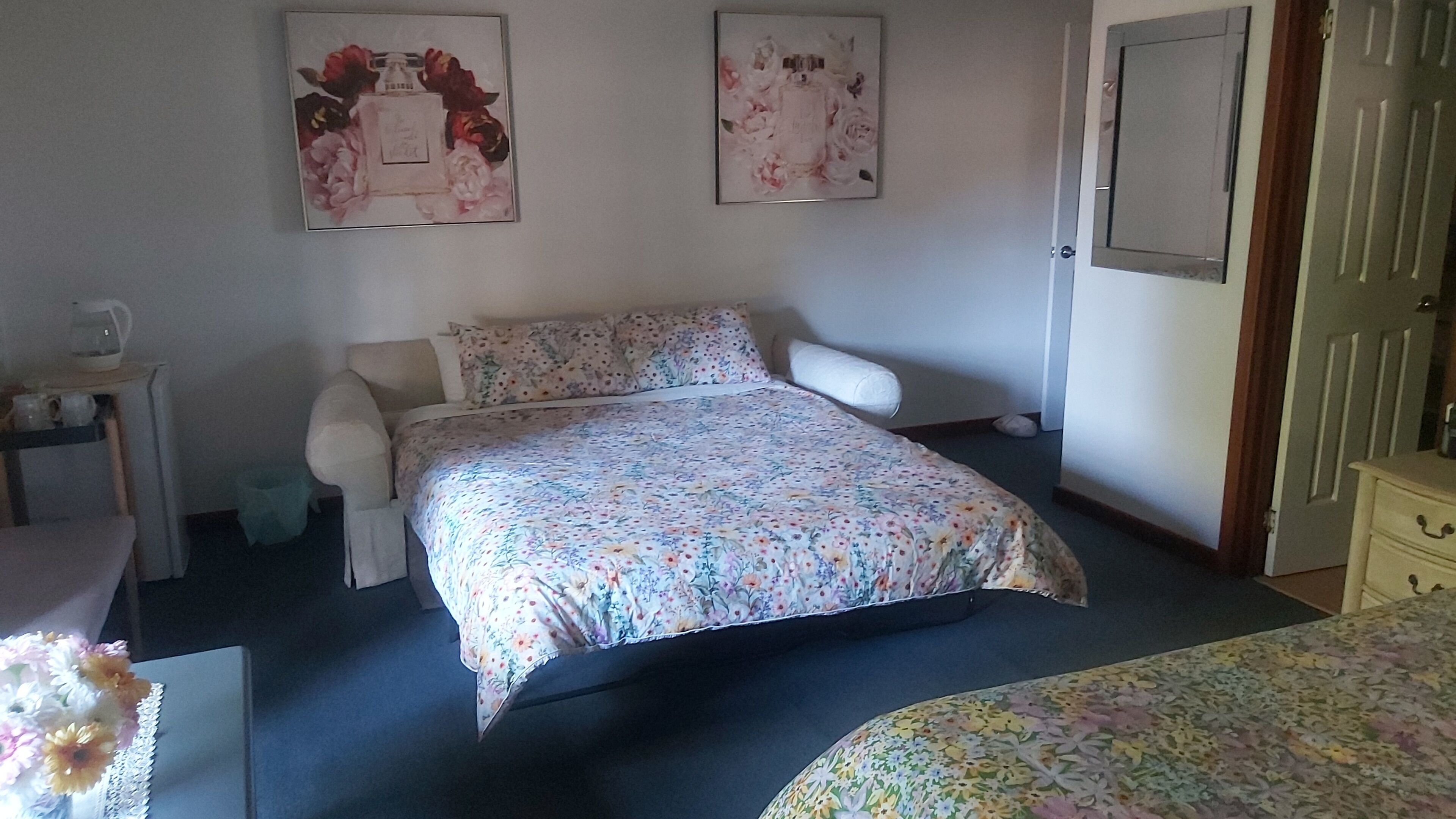 A bedroom with a floral bedspread, a bedside table with a kettle, and two framed floral posters on the wall.