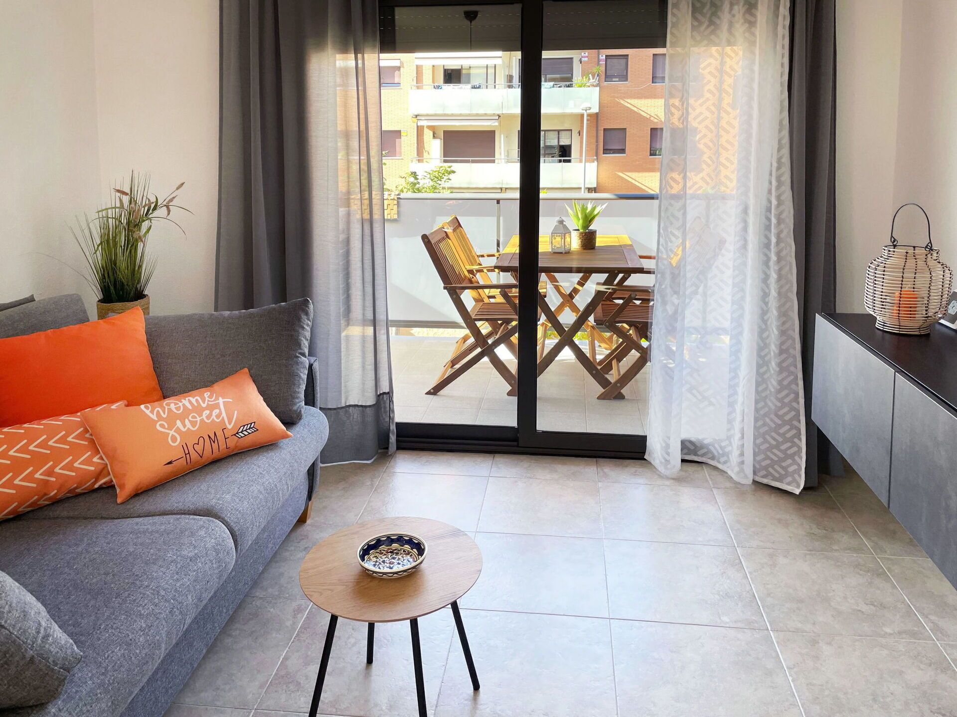 Ein modernes Wohnzimmer mit einer grauen Couch, orangefarbenen Kissen, einem kleinen Holztisch und einem Balkon mit einem Essplatz.