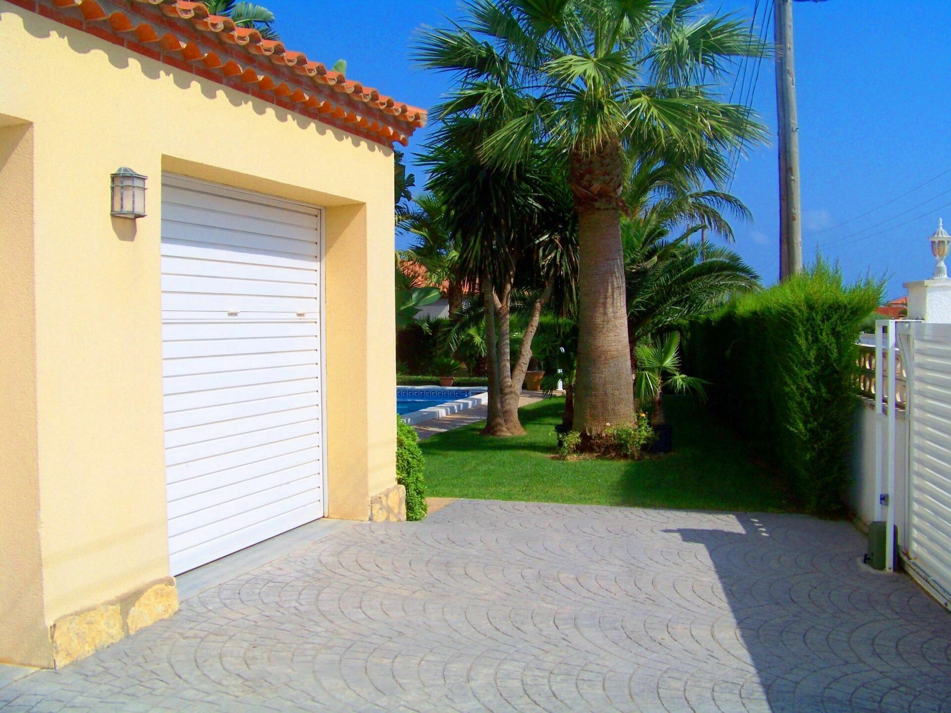 A building with a white garage door, a tiled roof, and a paved driveway leading to a well-maintained lawn with palm trees.