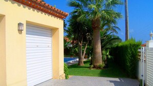 A building with a white garage door, a tiled roof, and a paved driveway leading to a well-maintained lawn with palm trees. - Beautiful private villa for 14 people with private pool, WIFI, TV, patio and pets allowed (Miami Platja)