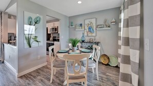 A dining area with a round table, chairs, and a potted plant. There is a kitchen in the background with a microwave and shelves. - 2nd Floor, Shared Pool Hot Tub, Near Marina, Pickleball courts (Port Aransas)