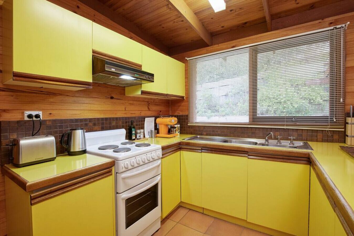 Une cuisine de taille réduite avec des meubles de couleur jaune, une cuisinière, un évier et une fenêtre équipée de stores.