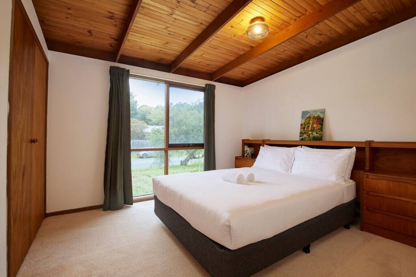 A bedroom with a bed, wooden headboard, a nightstand, a painting, and a view through a window.