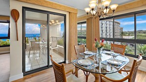 A dining area with a glass table, chairs, and a chandelier. There is a view of the ocean through large windows. - MRR Presents Kaanapali Ali'i 282 -  Remodeled 8th Floor Corner 2BR w/ Partial Ocean AND Mountain Views! (Lahaina)