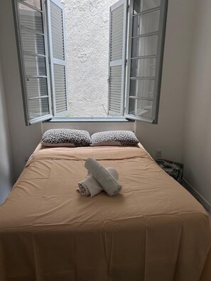 Ett sovrum med en säng, två kuddar, en hoprullad handduk och ett fönster med fönsterluckor.
