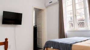 Un dormitorio con cama, televisión, equipo de aire acondicionado, ventana con cortinas y puerta que da a otra habitación.